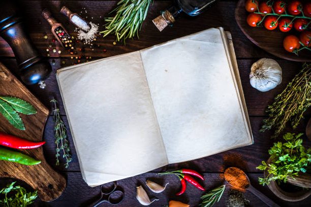 Top view of a vintage open cookbook surrounded by some spices and herbs like thyme, rosemary, laurel, pepper, garlic and chervil, and some kitchen utensils. The cookbook is at the center of the image and its pages are empty so you can use it as a useful copy space. Objects are on a rustic dark brown wooden table. 